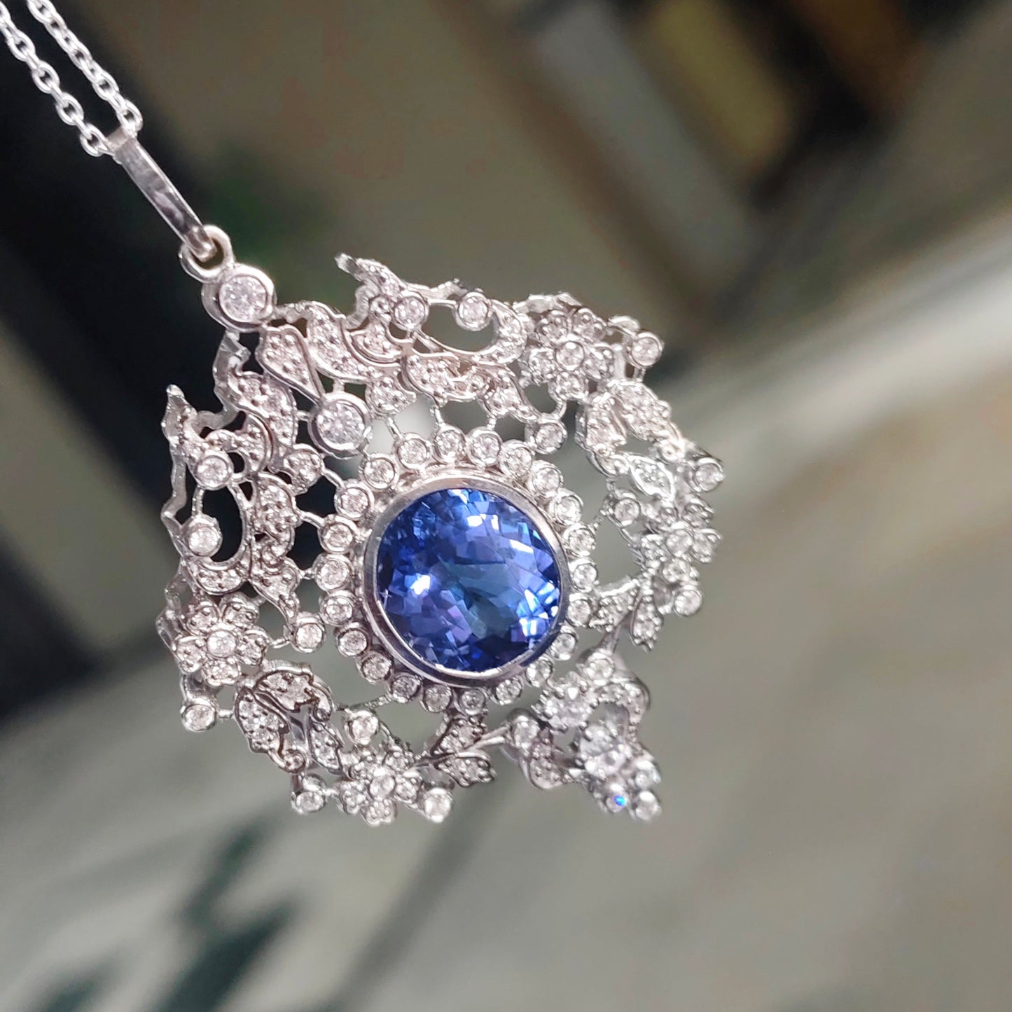 An ornate silver-toned pendant necklace featuring a central round blue gemstone surrounded by intricate filigree and small clear stones, hanging against a blurred indoor background.