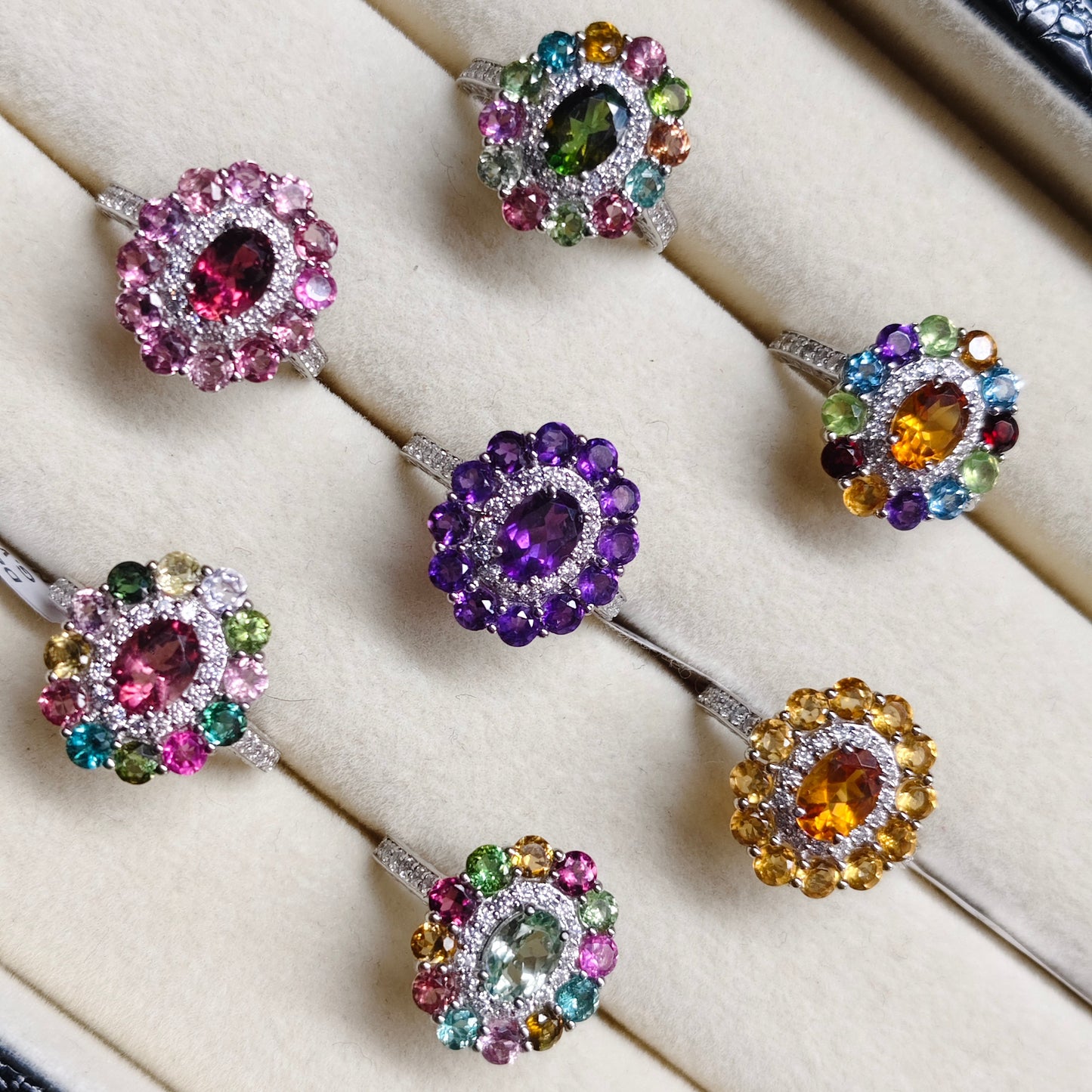 Six colorful gemstone halo rings displayed in a cream-colored jewelry box. The rings feature silver-toned settings with various colored gemstones including pink, green, purple, orange, and yellow, surrounded by clear accent stones.
