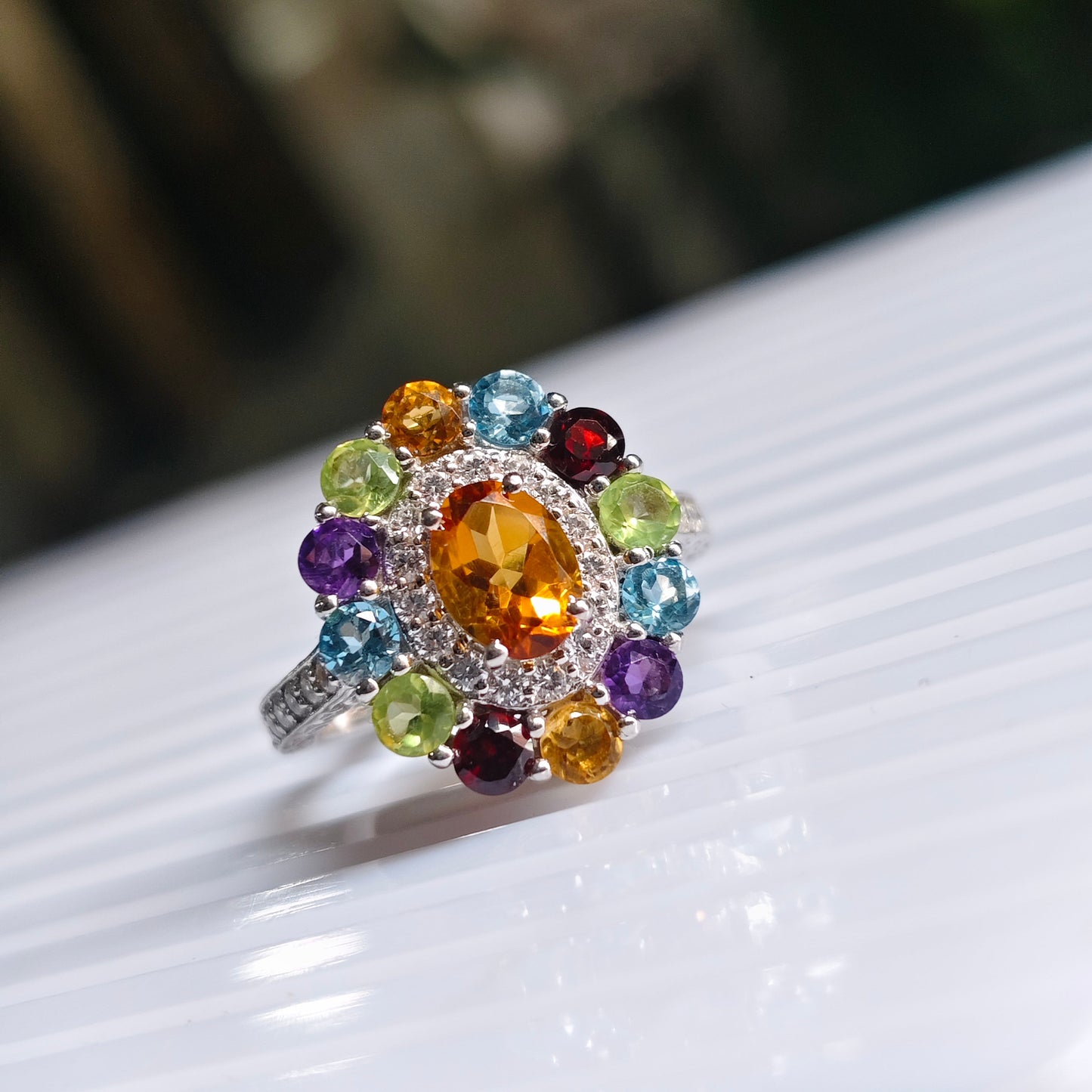 A silver-toned ring featuring a large oval orange center stone surrounded by a halo of small clear stones and a border of multicolored round gemstones, resting on a white ribbed surface.
