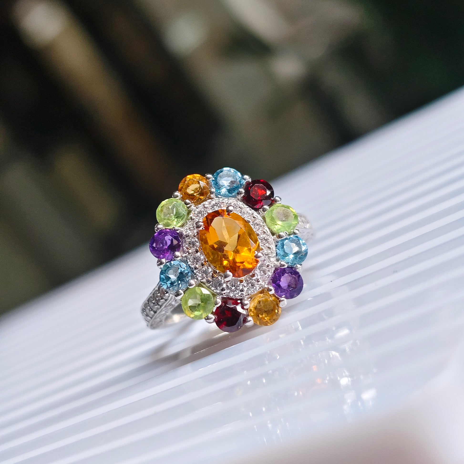 A colorful gemstone cluster ring with a central orange oval stone surrounded by multicolored round stones, set on a silver band, resting on a white ribbed surface.