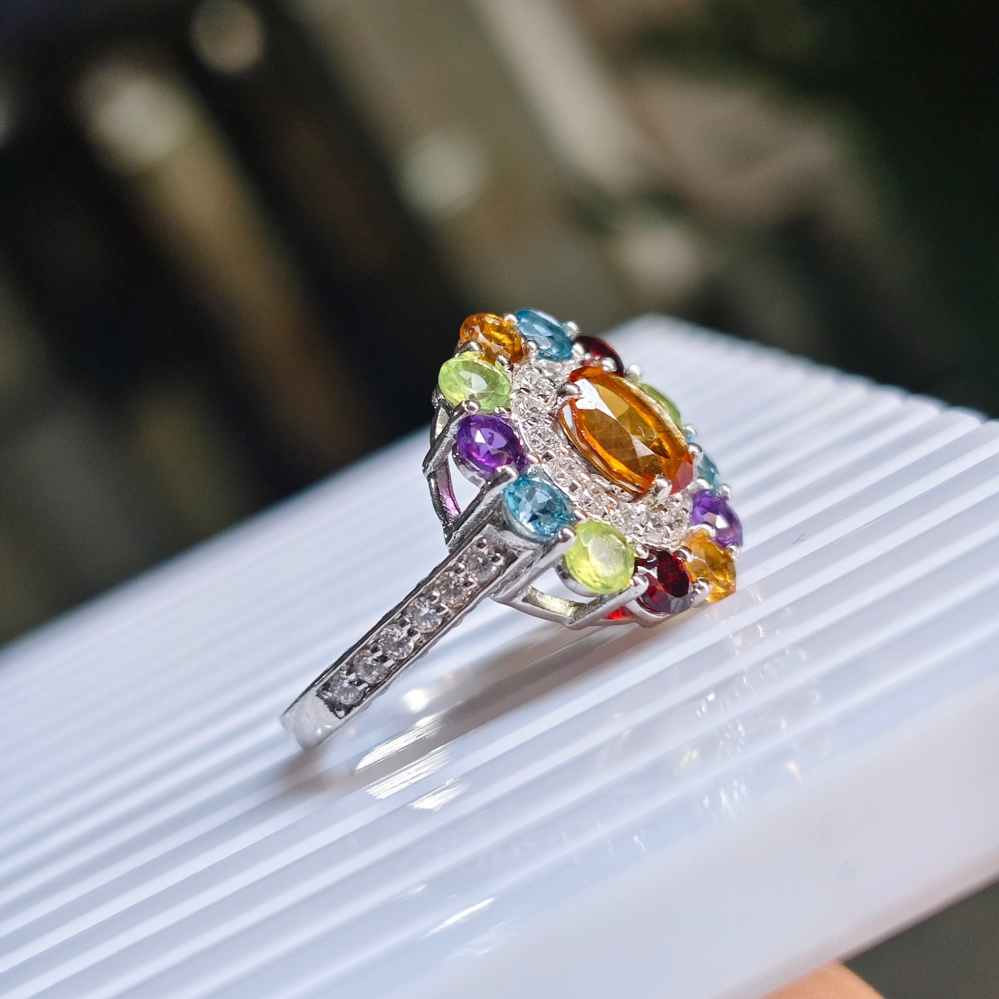 A silver ring with a colorful gemstone cluster, featuring a large oval orange center stone surrounded by smaller multicolored stones, resting on a white ribbed surface.
