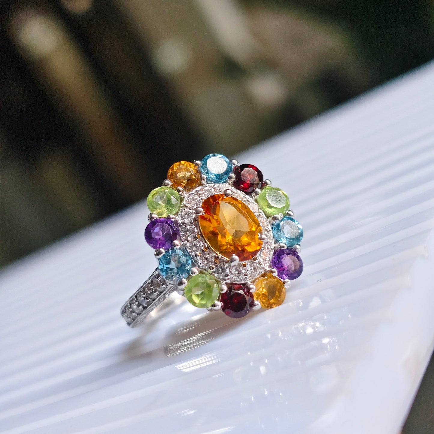 A silver ring with a large oval orange-yellow center stone and a surrounding halo of multicolored round gemstones, resting on a white ribbed surface.