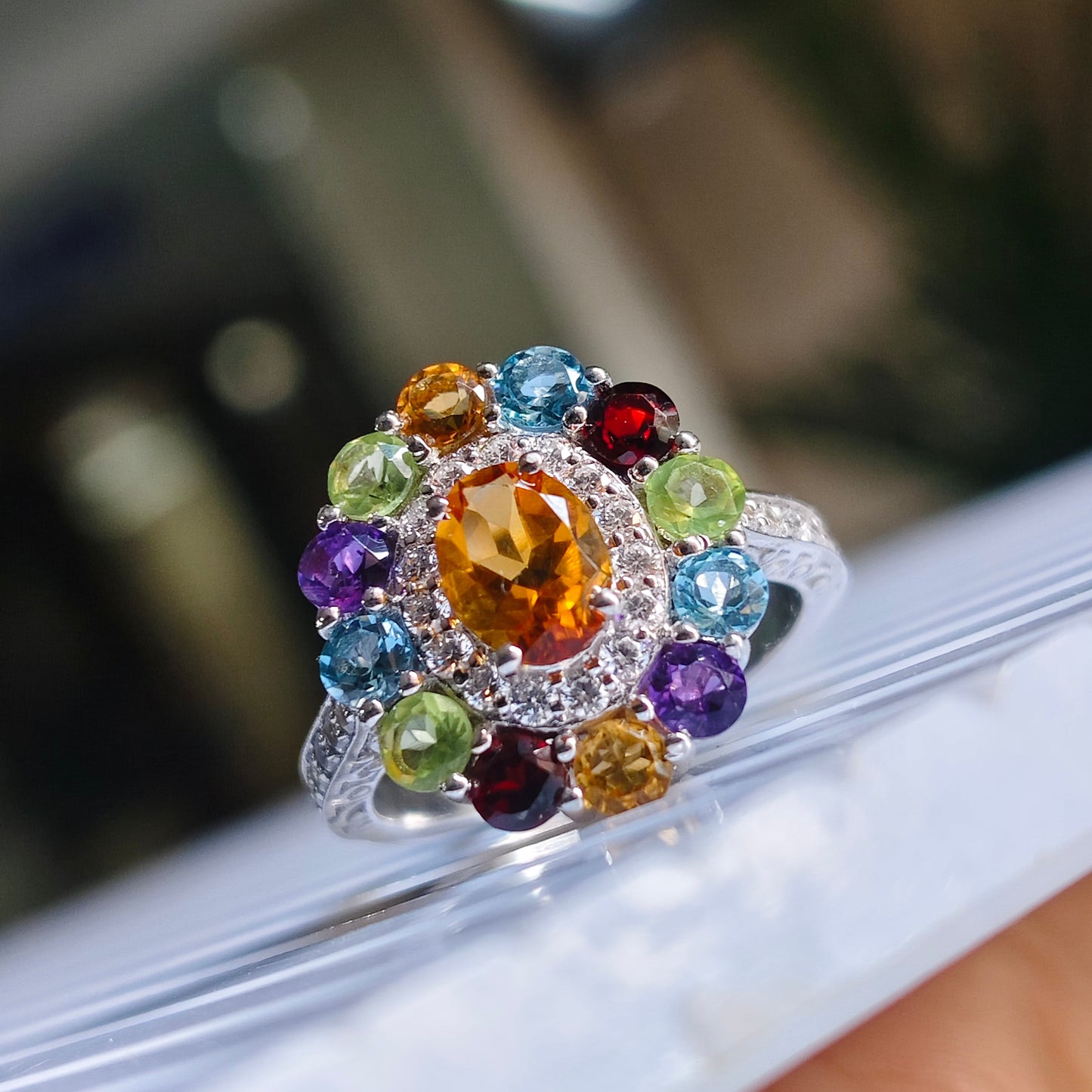 A silver-toned cluster ring with a large oval amber center stone and multiple round-cut gemstones in blue, green, purple, red, and yellow, shown close up on a white surface.