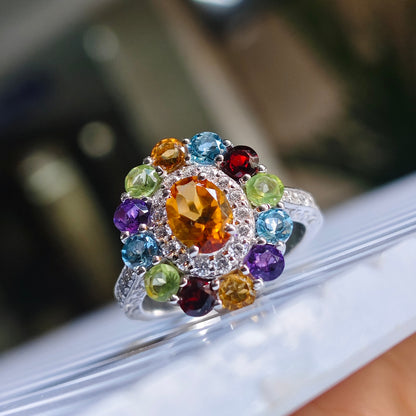 A silver-toned cluster ring with a large oval amber center stone and multiple round-cut gemstones in blue, green, purple, red, and yellow, shown close up on a white surface.