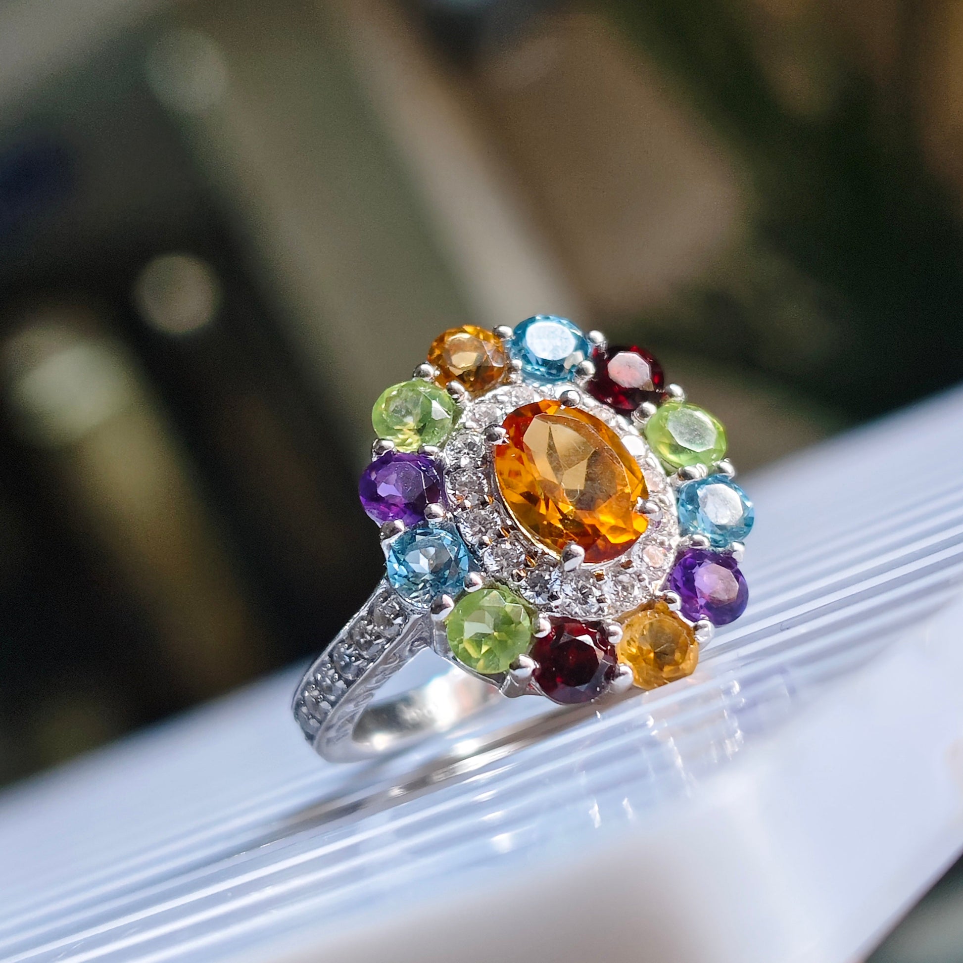 A silver-toned cluster ring with a large oval orange-yellow center stone surrounded by multiple round multicolored gemstones, shown resting on a white ribbed surface.