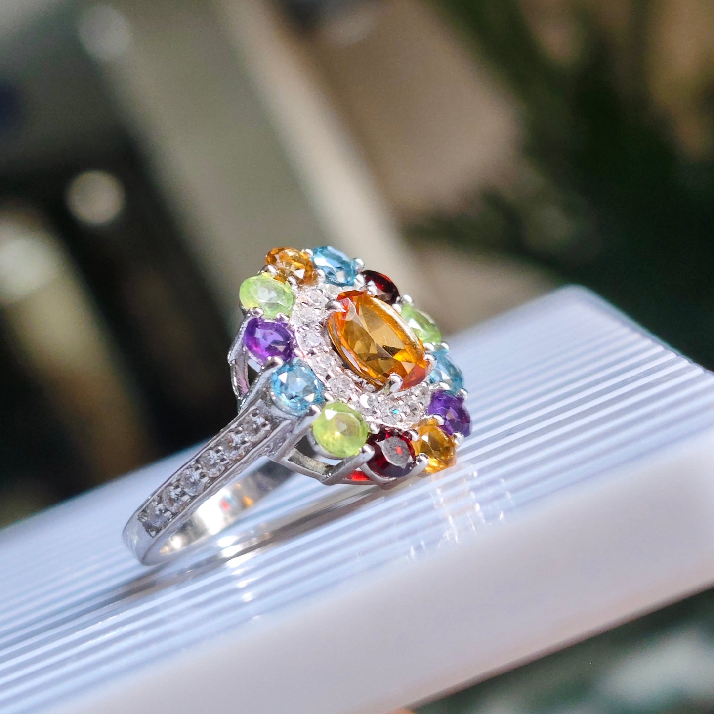 A silver ring with a colorful gemstone cluster, featuring a large oval orange center stone surrounded by smaller multicolored stones, resting on a white ribbed surface.