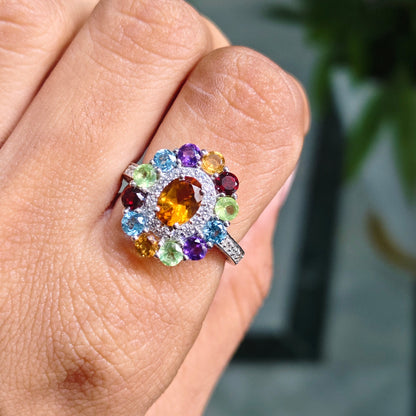 A silver-toned ring with a multicolor gemstone cluster, worn on a person's finger. The ring features a central oval yellow gemstone surrounded by various colored round stones in purple, blue, green, and orange, set in a silver-toned band.