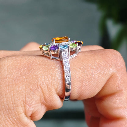 A silver-toned ring with a multi-colored gemstone cluster and a channel-set clear stone down the band, shown worn on a person's finger against a blurred gray background with a green plant.