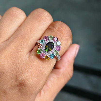 A close-up of a hand wearing a colorful gemstone ring. The ring features a central oval dark green stone surrounded by a halo of pink, blue, green, and yellow stones in a silver-toned setting.