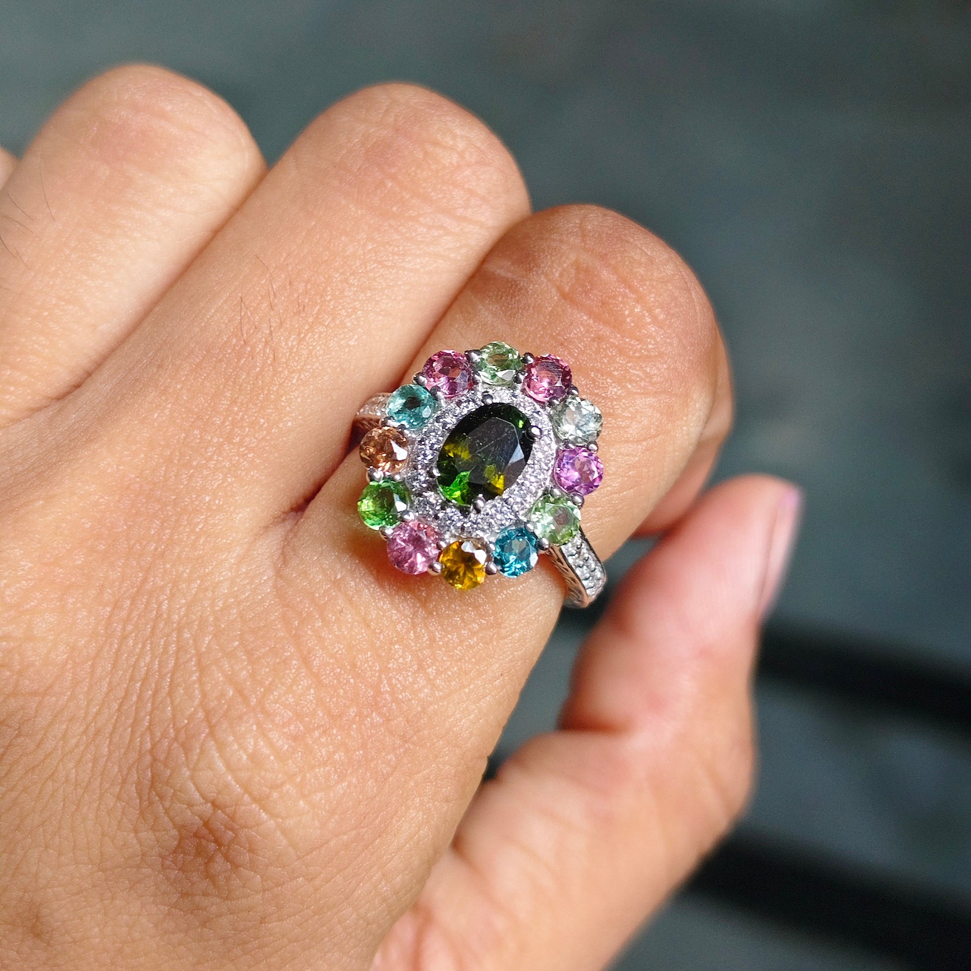 A close-up of a hand wearing a colorful ring with a central dark oval stone surrounded by multicolored gemstones in pink, green, blue, yellow, and orange on a silver-toned setting.