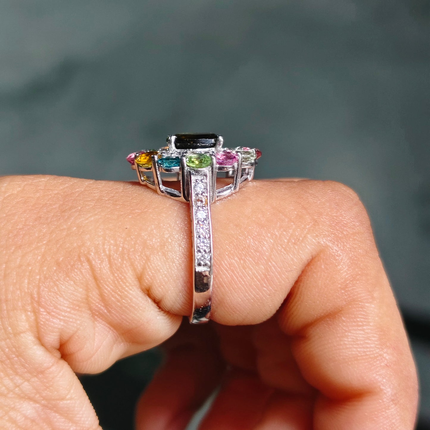 A silver-toned ring with a rectangular black center stone and small multicolored accent stones on either side, shown worn on a person's finger against a dark gray blurred background.
