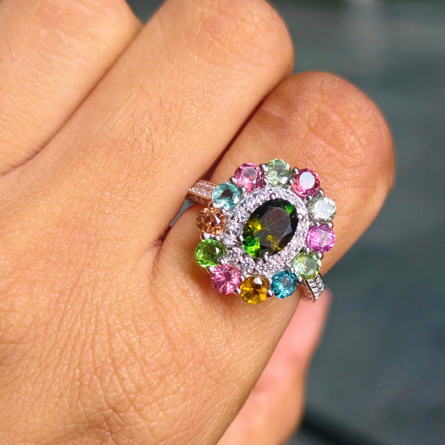 A close-up of a hand wearing a colorful gemstone ring. The ring features a silver-toned band and a cluster of round-cut gemstones in pink, blue, green, yellow, and purple around a central dark oval stone.