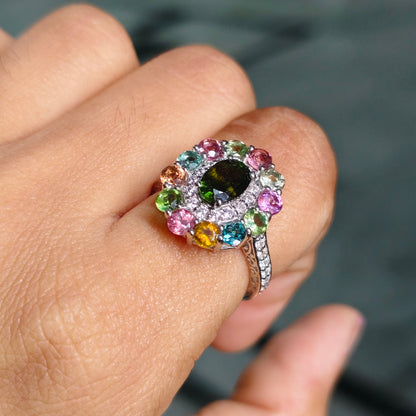 A close-up of a hand wearing a colorful gemstone ring. The ring features a large central dark green oval stone surrounded by a halo of small multicolored round-cut gemstones in pink, blue, yellow, and green, set in a silver-colored band.