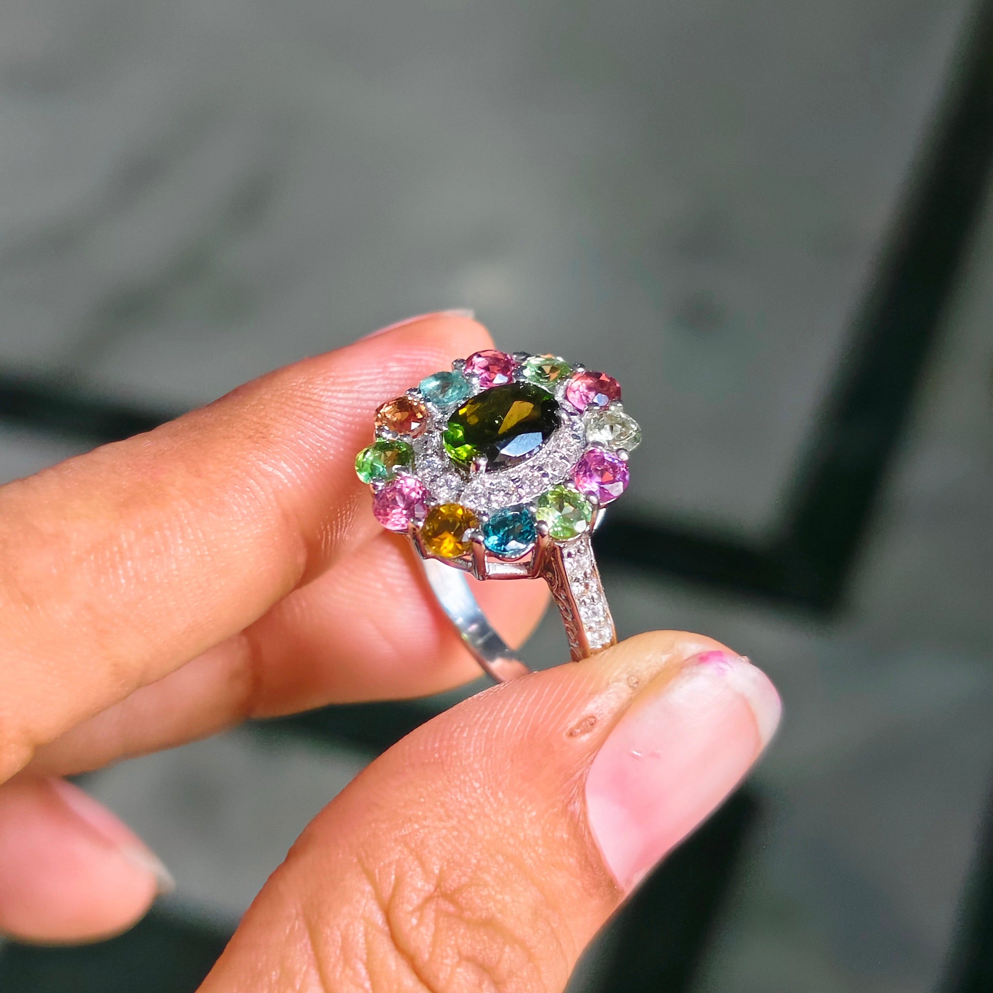 A close-up of a hand holding a silver ring with a colorful gemstone cluster. The ring features a central olive-green heart-shaped gemstone surrounded by a halo of pink, blue, green, and yellow accent stones.