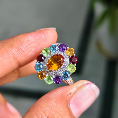 A close-up of a hand holding a silver ring with a colorful gemstone cluster. The ring features a large central round yellow gemstone surrounded by a halo of smaller multicolored gemstones in green, blue, purple, red, and orange, set in a silver-toned band.