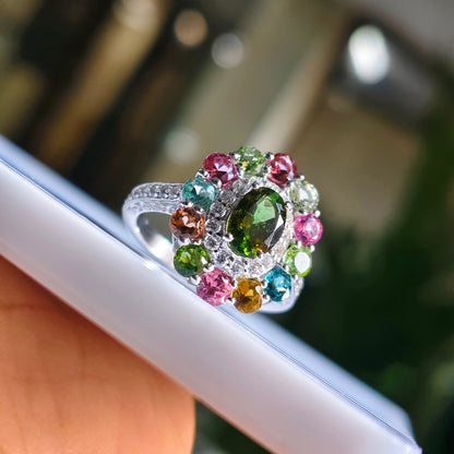 A silver ring with a large oval green gemstone surrounded by a colorful halo of pink, green, blue, and orange accent stones, held by a hand near a white rectangular surface.