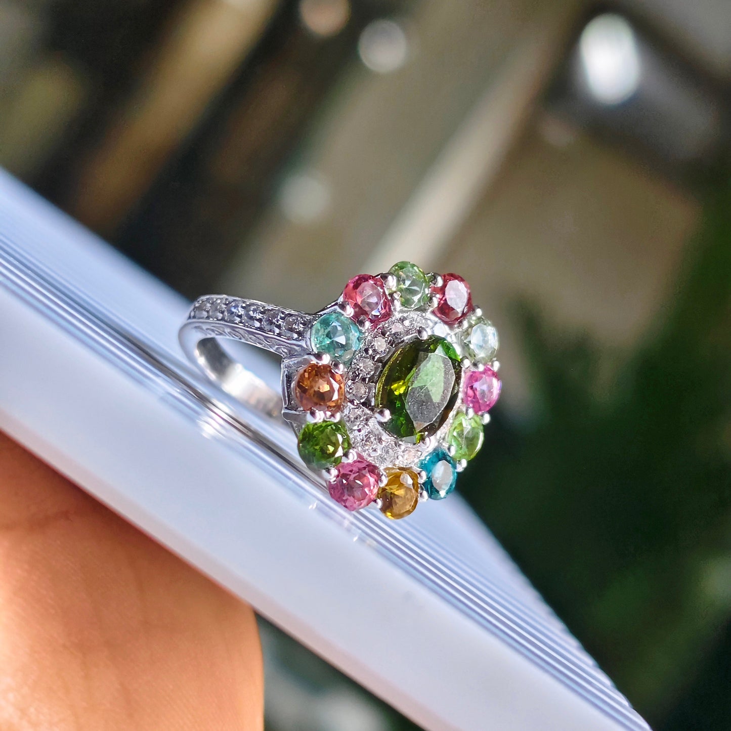 A silver ring with a colorful gemstone cluster, held by a hand against a blurred outdoor background.