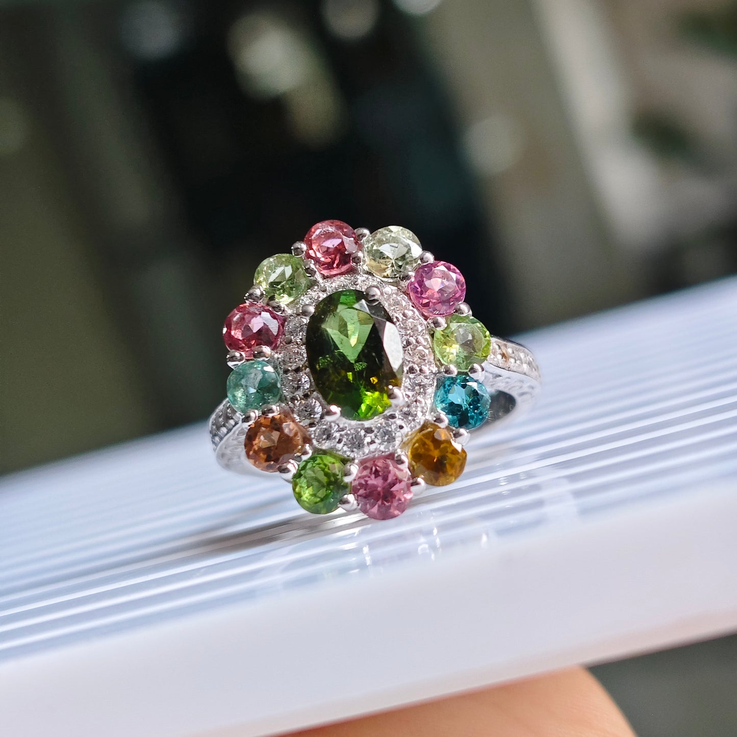 A silver-toned cluster ring with a large green oval center stone and multiple smaller multicolored round stones, shown resting on a white ribbed surface with a hand holding the bottom edge.