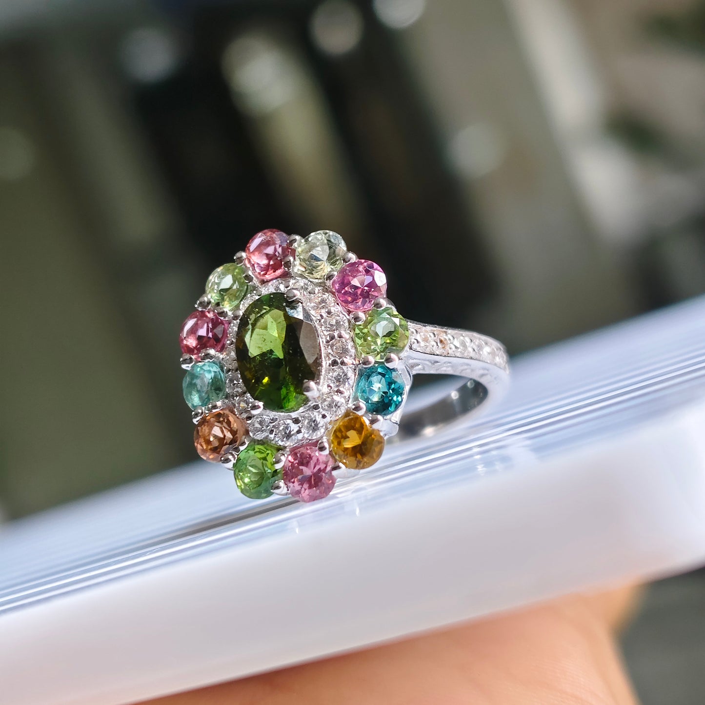 A silver ring with a colorful gemstone cluster, featuring a green oval center stone surrounded by pink, blue, green, and orange round stones, resting on a white surface held by a hand.