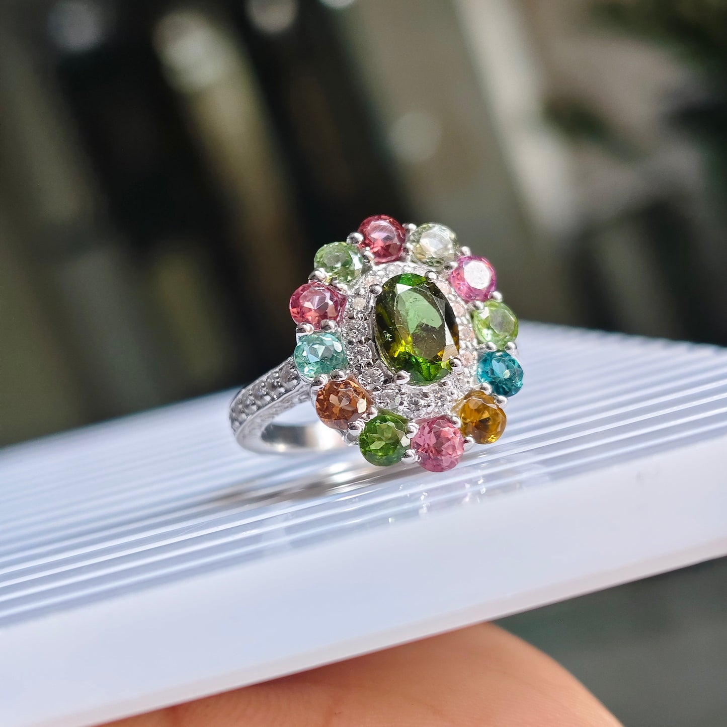 A silver ring with a colorful gemstone cluster, featuring a central green oval stone surrounded by pink, blue, green, and yellow stones, resting on a white ribbed surface held by a hand.