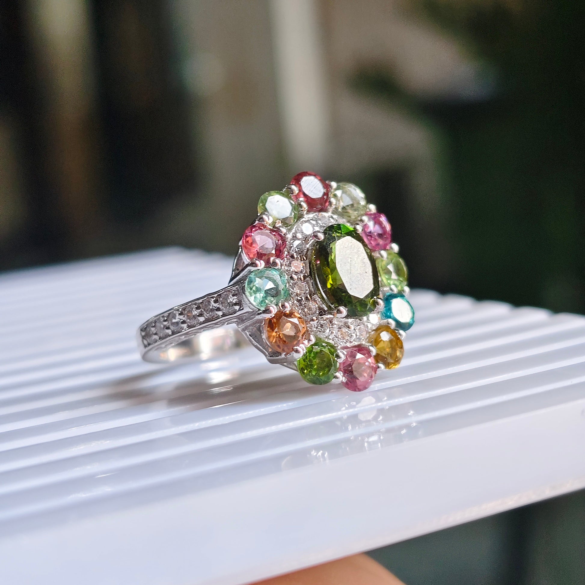 A silver-toned ring with a colorful gemstone cluster, featuring a central green oval stone surrounded by pink, blue, green, and orange stones, resting on a white ribbed surface.