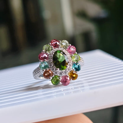 A silver-toned cluster ring with a large green oval center stone and multiple smaller multicolored gemstones, shown resting on a white ribbed surface held by a hand.