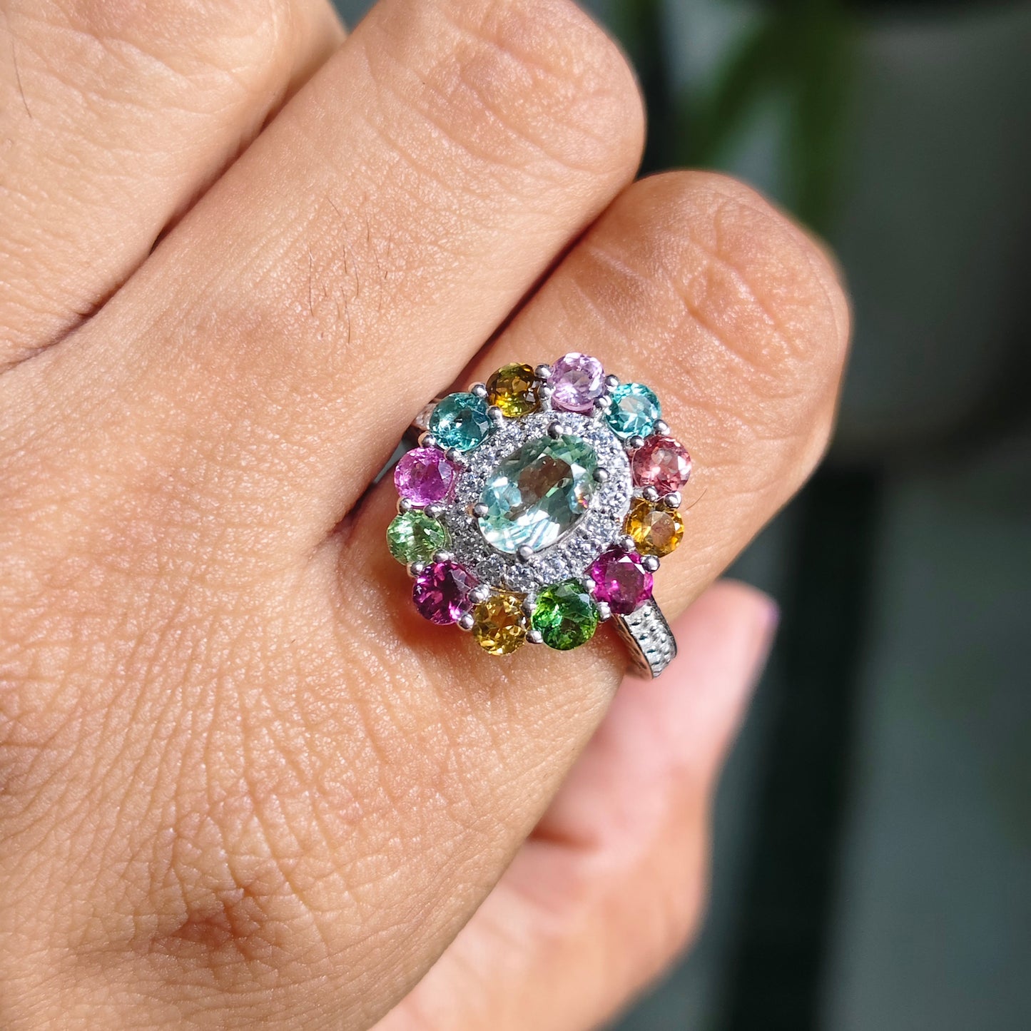 A close-up of a hand wearing a silver-toned ring with a large clear center stone and a surrounding halo of multicolored gemstones, including pink, blue, green, yellow, and orange.