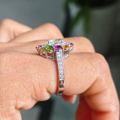A close-up of a hand wearing a silver-toned ring with a large clear center stone and small colorful side stones in pink, green, and yellow.