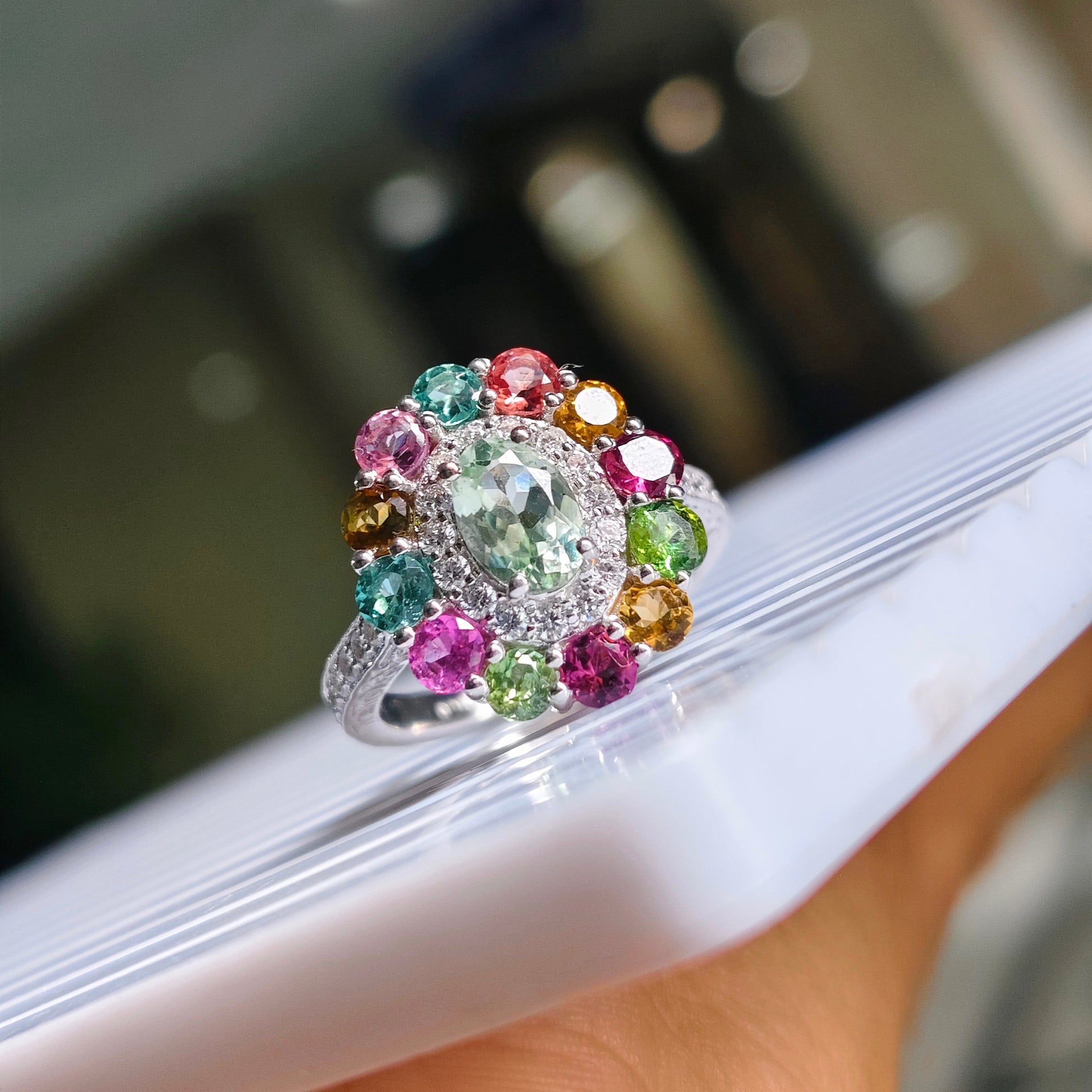 A silver-toned cluster ring with a large light green center stone and multiple surrounding multicolored gemstones, shown resting on a white surface with a blurred indoor background.