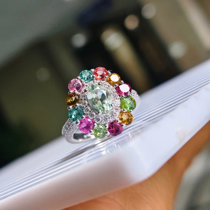 A silver-toned cluster ring with a large light green center stone and multiple surrounding multicolored gemstones, shown resting on a white surface with a blurred indoor background.