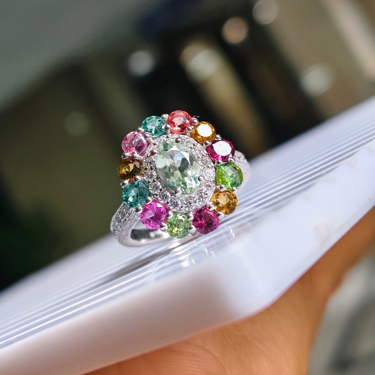 A silver-toned cluster ring with a large light green center stone and multiple surrounding multicolored gemstones, shown resting on a white surface with a blurred indoor background.