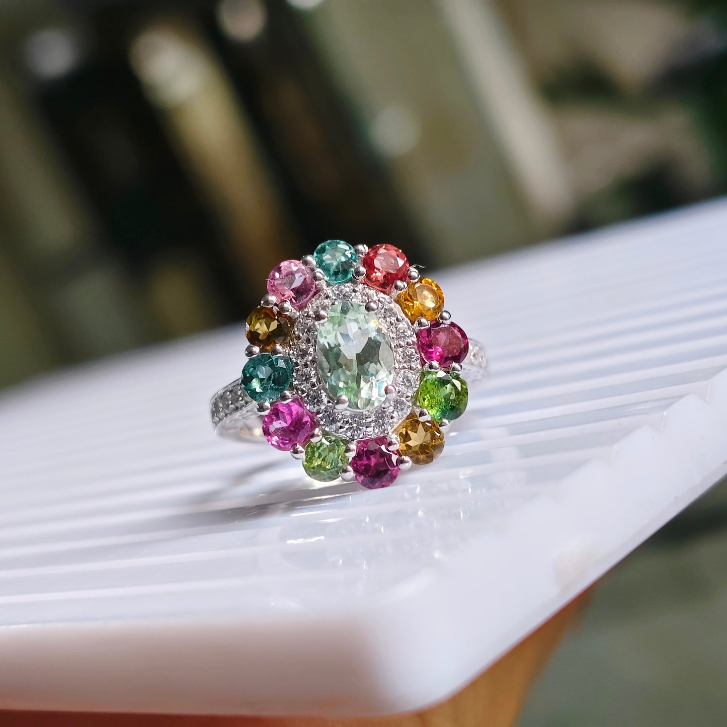 A silver ring with a large oval green center stone and a halo of colorful round gemstones in pink, green, yellow, and orange, resting on a white marble surface.