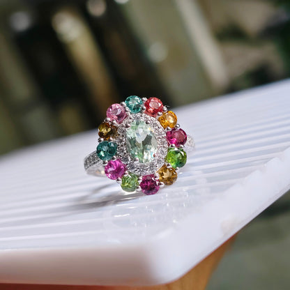 A silver ring with a large oval green center stone and a halo of colorful round gemstones in pink, green, yellow, and orange, resting on a white marble surface.