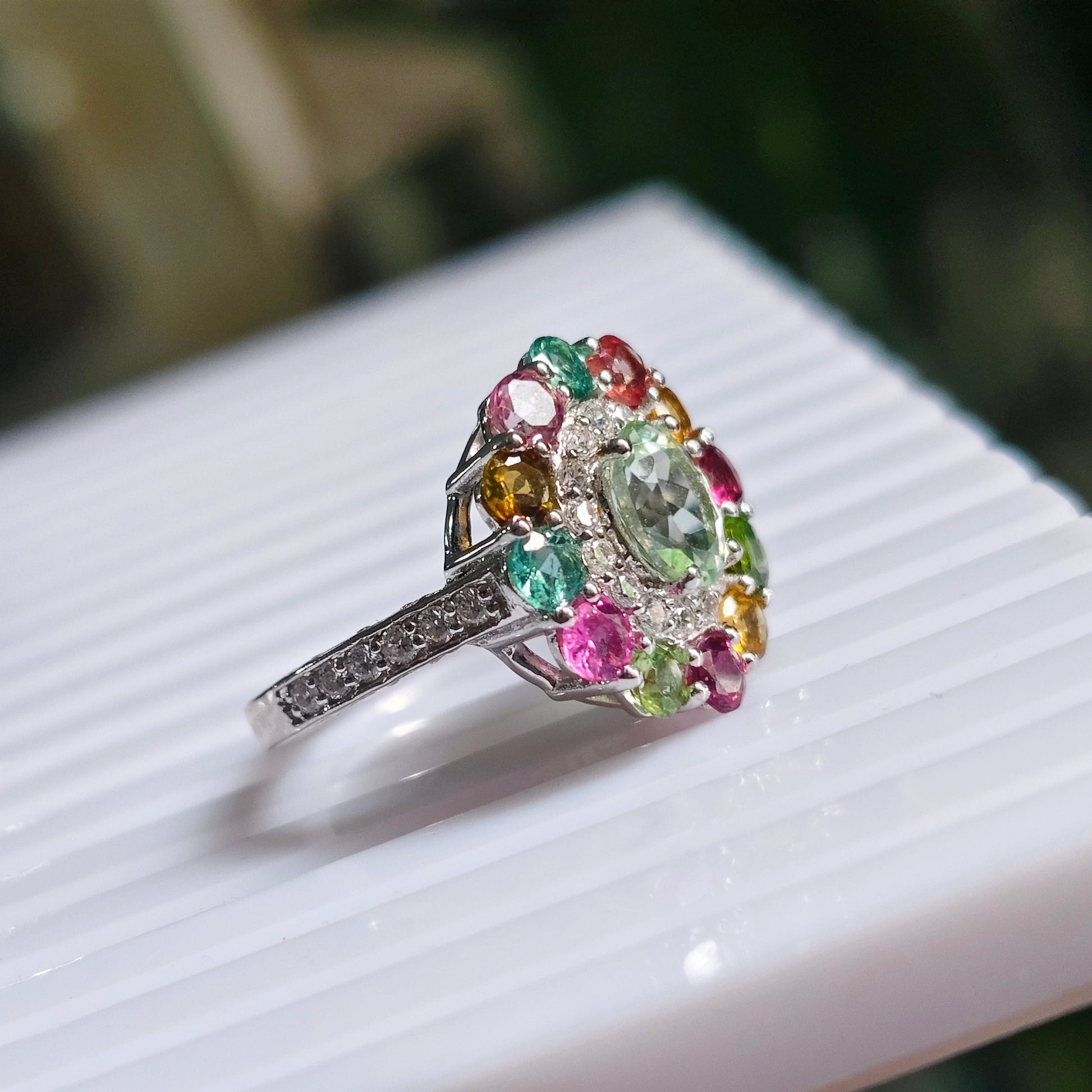 A silver-toned ring with a large, colorful floral cluster of round-cut gemstones in pink, green, yellow, and blue, resting on a white ribbed surface.