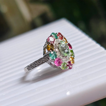 A silver-toned ring with a large, colorful floral cluster of round-cut gemstones in pink, green, yellow, and blue, resting on a white ribbed surface.
