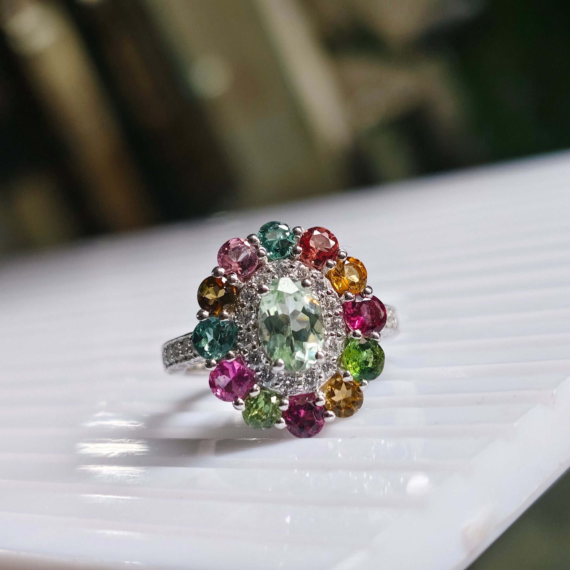 A silver-toned cluster ring featuring a central oval green gemstone surrounded by a halo of multicolored round gemstones, resting on a white ribbed surface with a blurred dark background.