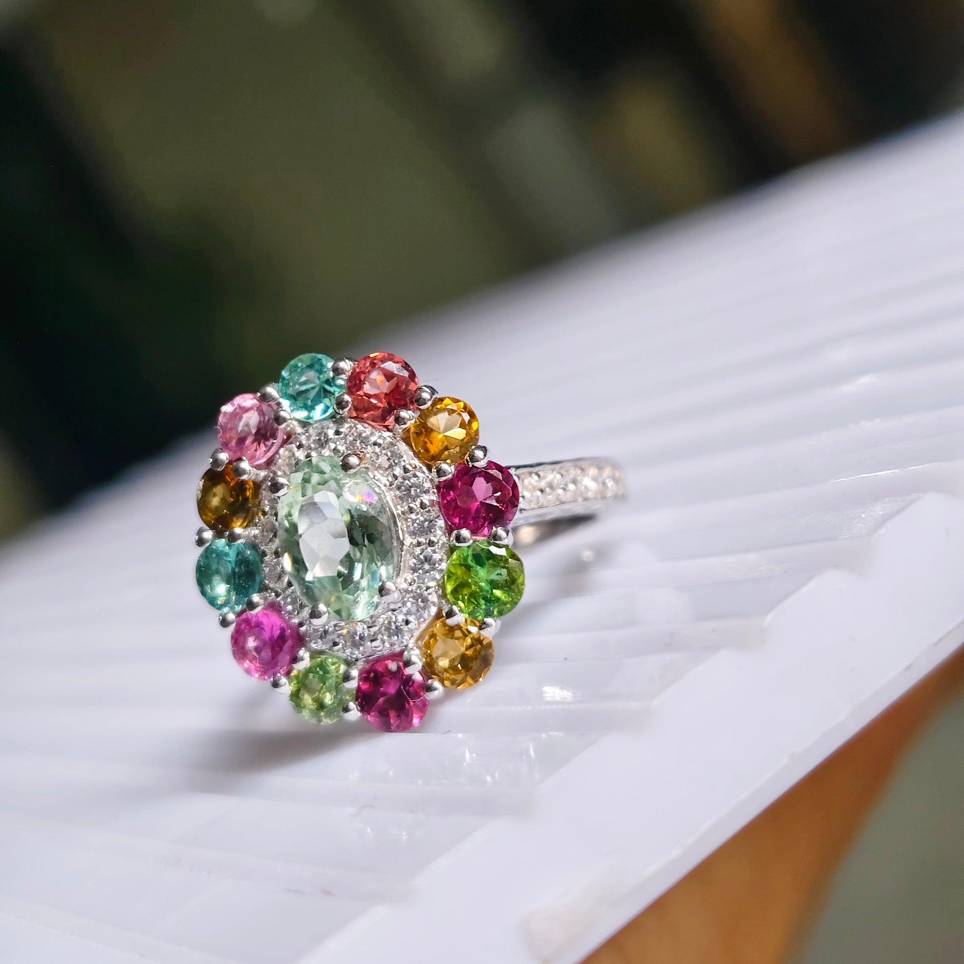 A silver ring with a colorful gemstone cluster, featuring a large oval-cut light green center stone surrounded by smaller multicolored stones in pink, red, yellow, and green, resting on a white ribbed surface.