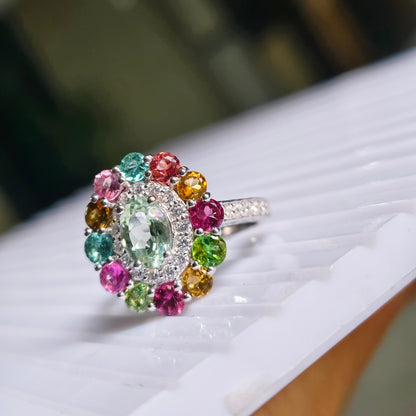 A silver ring with a colorful gemstone cluster, featuring a large oval-cut light green center stone surrounded by smaller multicolored stones in pink, red, yellow, and green, resting on a white ribbed surface.