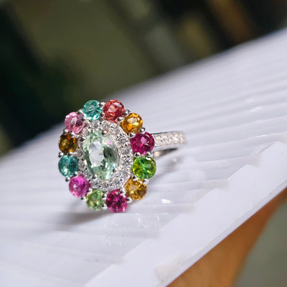 A silver ring with a colorful gemstone cluster, featuring a large oval-cut light green center stone surrounded by smaller multicolored stones in pink, red, yellow, and green, resting on a white ribbed surface.