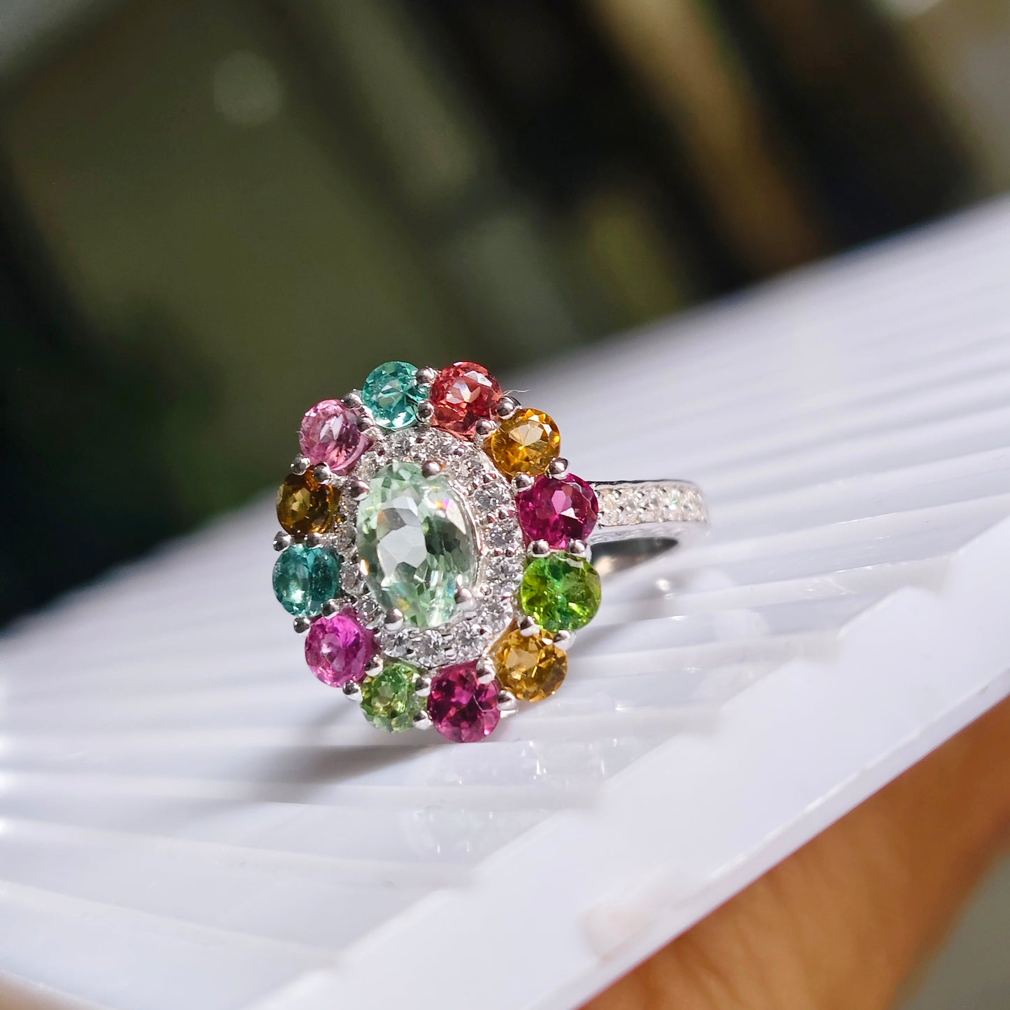 A multicolor gemstone cluster ring with a central marquise-cut green stone surrounded by colorful round-cut stones in pink, blue, green, yellow, and orange, set on a silver-toned band and resting on a white textured surface.