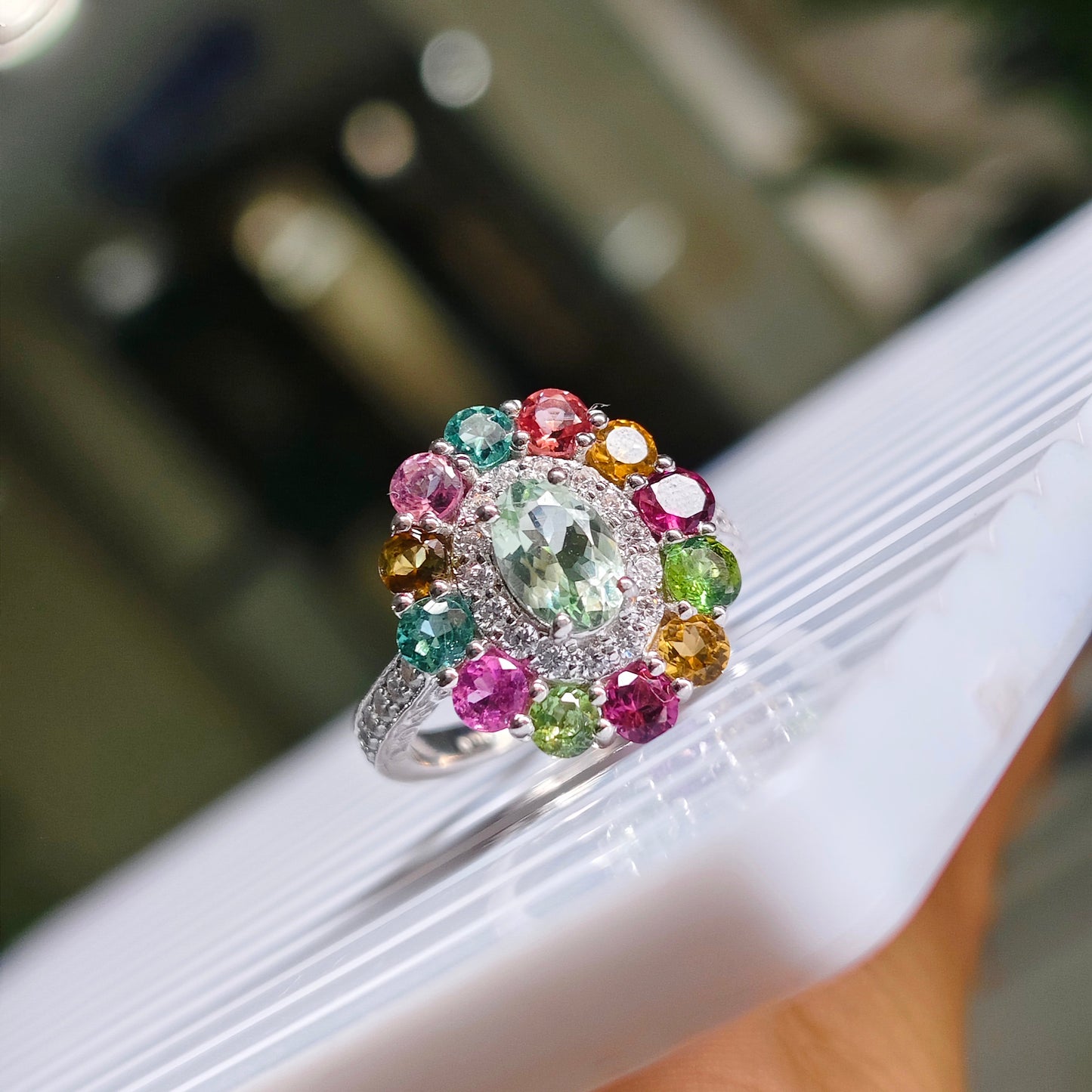 A silver-toned cluster ring with a large oval clear center stone and surrounding multicolored round gemstones, shown resting on a white ribbed surface with a hand holding it. The background is heavily blurred.