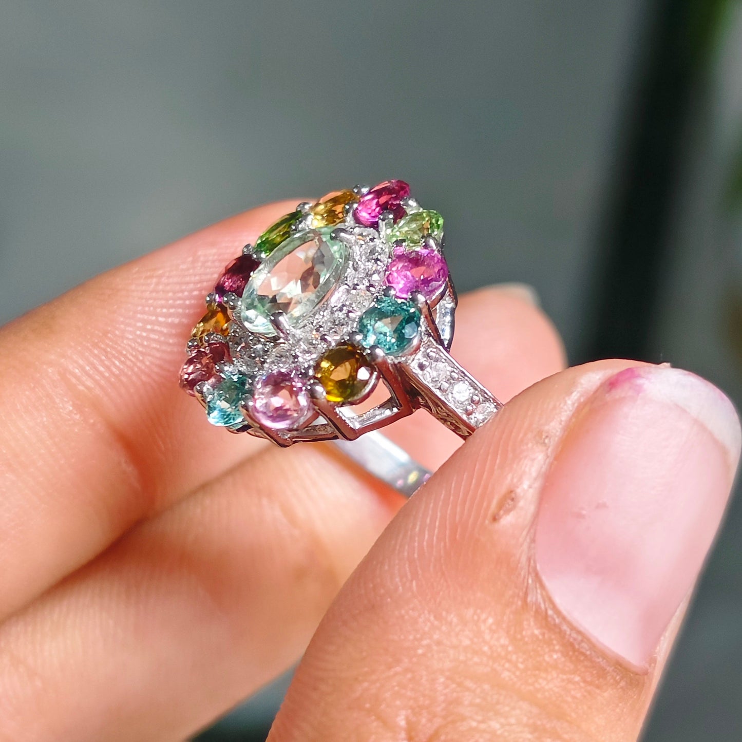 A close-up of a hand holding a colorful gemstone ring. The ring features a large clear central stone surrounded by small pink, green, and yellow accent stones in a silver-toned setting.