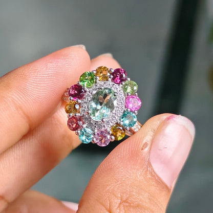 A close-up of a hand holding a colorful gemstone ring. The ring features a central clear round stone surrounded by a halo of small, multi-colored stones in pink, green, blue, yellow, and orange, set in a silver-toned metal.