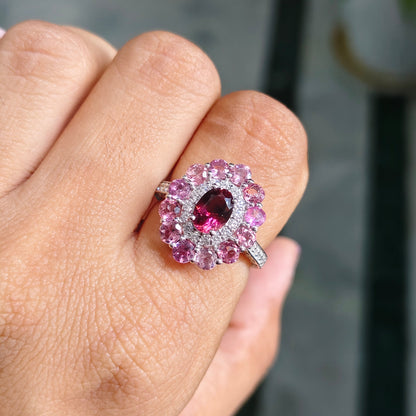 A close-up of a hand wearing a silver-toned ring featuring a central marquise-cut deep pink gemstone surrounded by a double halo of round pink stones and small clear accent stones.