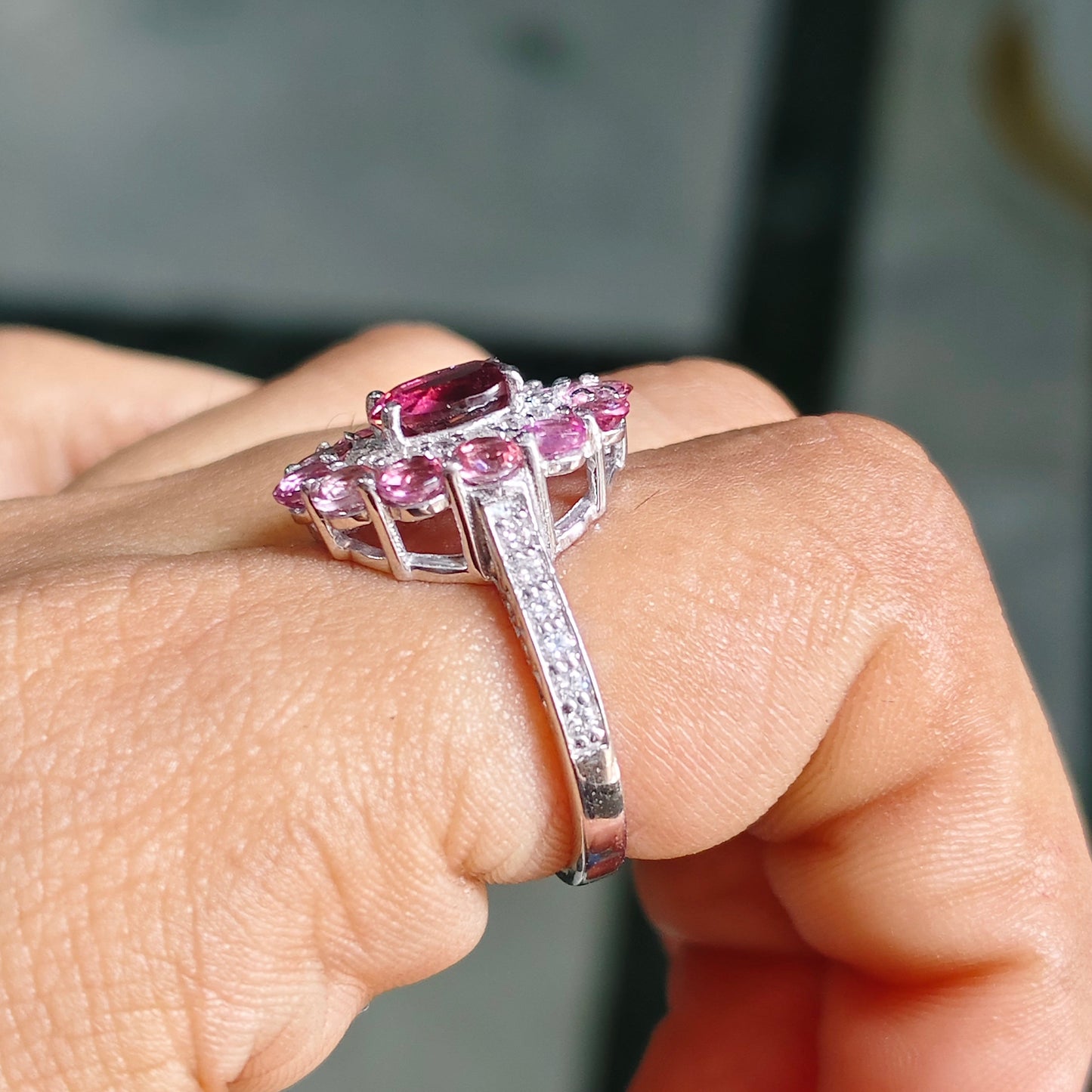 A close-up of a hand wearing a silver-toned ring with a large rectangular pink gemstone and smaller pink stones in a halo setting, with a pavé band.