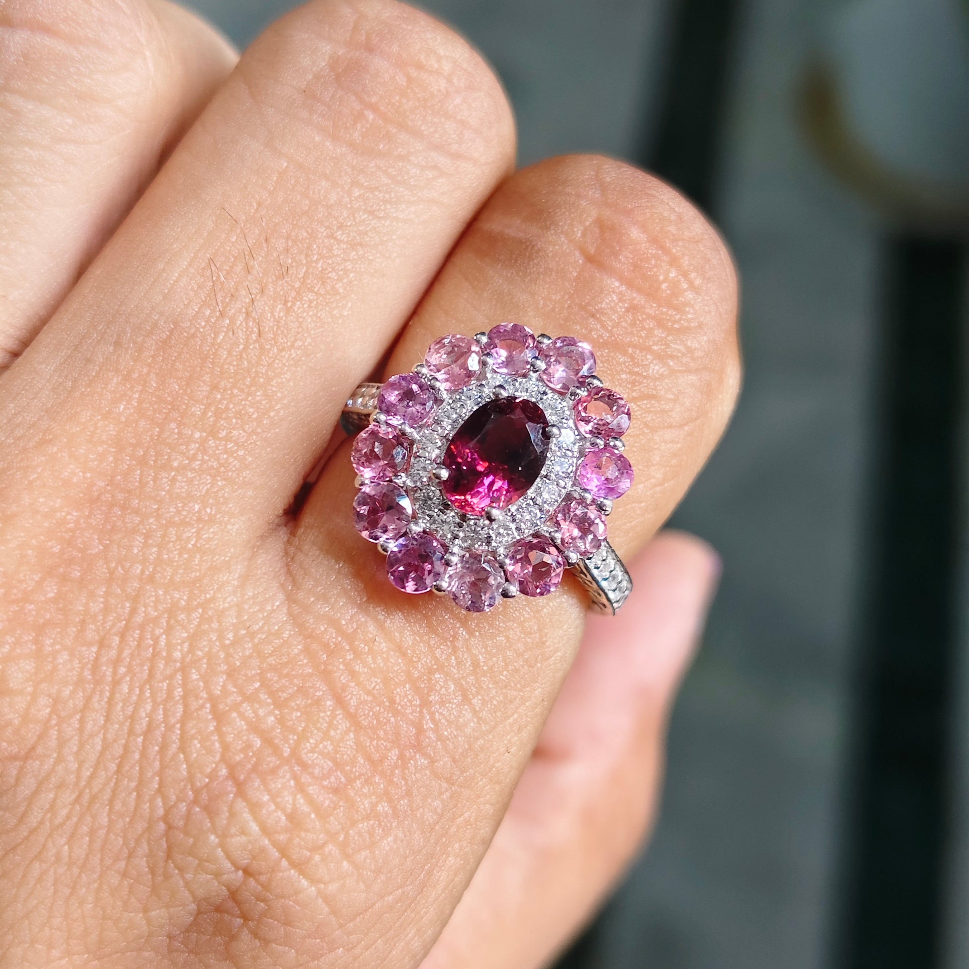 A close-up of a hand wearing a silver ring with a central marquise-cut pink gemstone surrounded by a halo of round pink and clear stones.