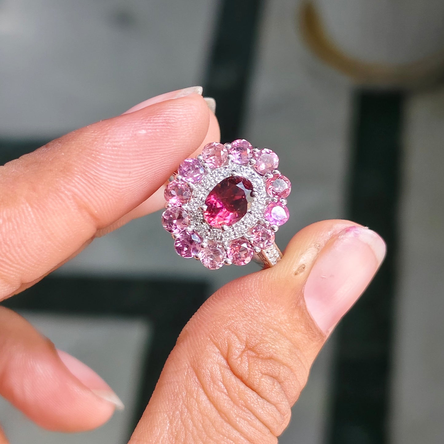 A hand holding a pink gemstone ring with a halo of small clear stones, photographed close-up against a blurred indoor background.