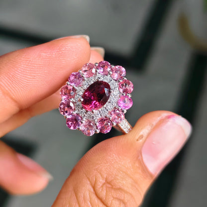 A close-up of a hand holding a silver ring featuring a central oval pink gemstone surrounded by a halo of round pink and clear stones.