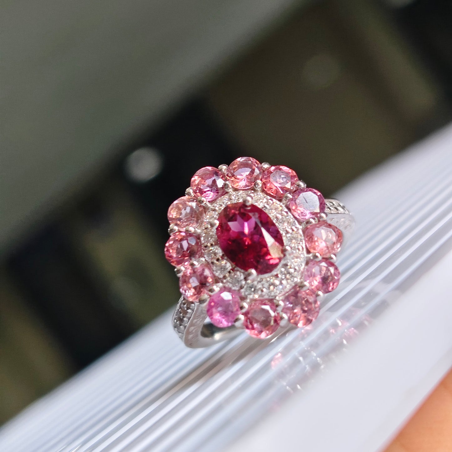 A close-up of a silver-toned ring featuring a central oval deep pink gemstone surrounded by a halo of round pink stones, displayed on a white ribbed surface.
