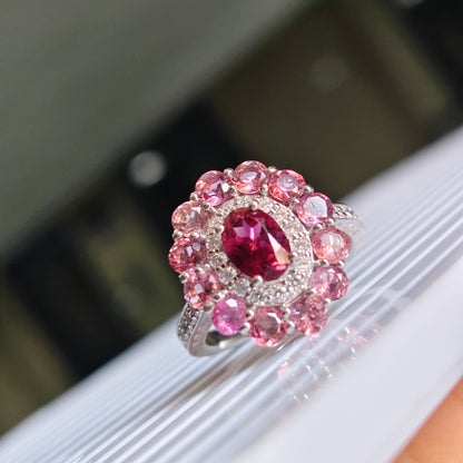 A close-up of a silver-toned ring featuring a central oval deep pink gemstone surrounded by a halo of round pink stones, displayed on a white ribbed surface.